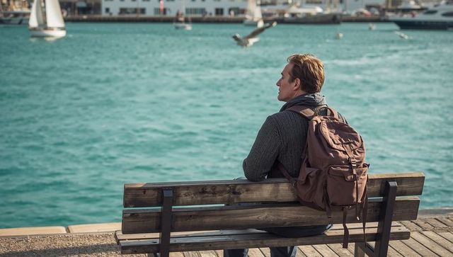 Solo traveler sitting on harbor bench gazing at sailboats, backpack on pier, turquoise sea
