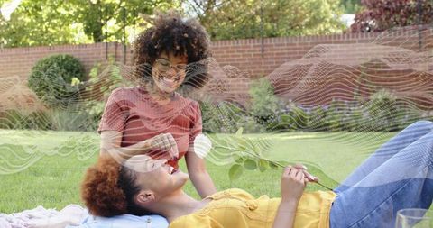 Smiling friends sharing picnic blanket and white flower, relaxing on sunlit backyard lawn