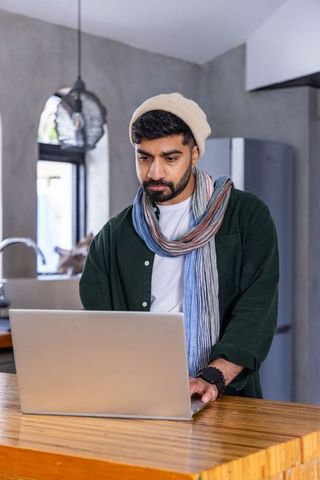 Young asian man working at kitchen island with laptop, beanie and scarf, remote work