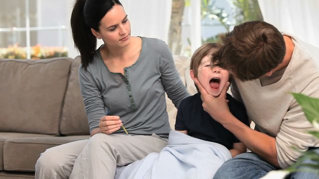 Parents are showing attention and care to their sick child on the sofa in a cozy living room. A loving mother holds a thermometer while the father checks the child's throat. The scene is perfect for topics about family health, parenting advice, childhood illness, and home healthcare solutions. It highlights compassion and nurturing within a family setting.