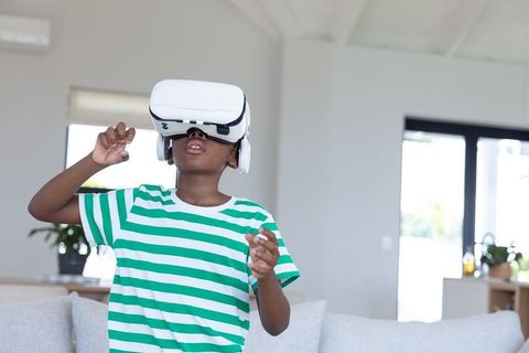 African american boy wearing vr headset exploring virtual world in bright living room