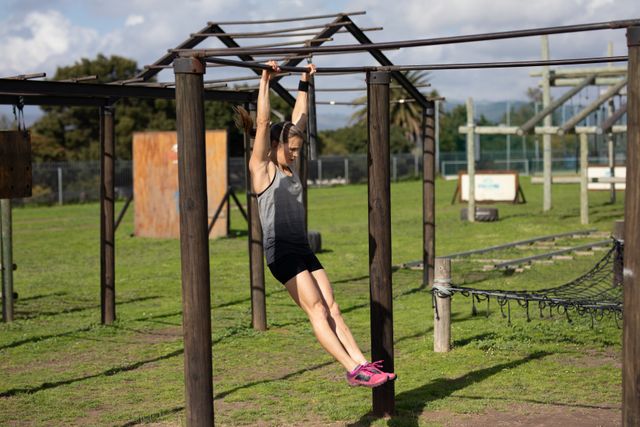 Side view of a Caucasian woman hanging from a bar, doing pull ups on a frame at an outdoor gym during a bootcamp training session, with other outdoor gym equipment in the background