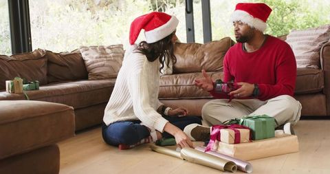 Couple wrapping holiday presents on cozy living room floor wearing santa hats