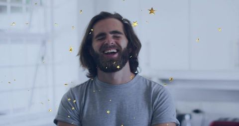 Laughing bearded man in gray t-shirt folding arms in sunlit minimalist kitchen with gold star confet