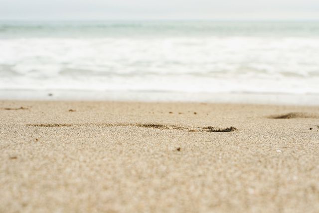 Picture captures close-up view of footprints in sand on beach with ocean waves in background. Can be used for themes such as travel, vacation, relaxation, beach activities, serenity, and nature's beauty. Useful for advertisements, travel articles, coastal property promotions, or summer-related marketing materials.