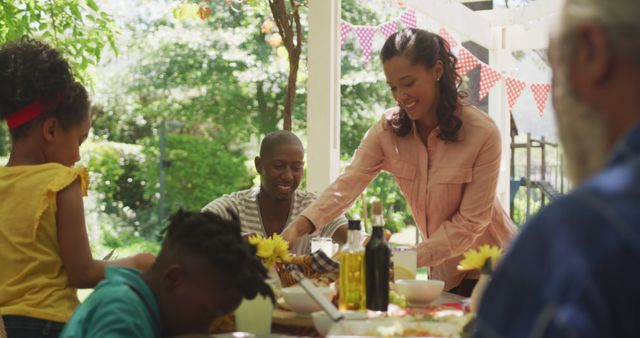 Family enjoying meal outdoors in a sunny garden. Parents and children sharing food and smiles around a dining table with festive bunting decorations. Perfect for themes of family bonding, festive celebrations, and summer gatherings.