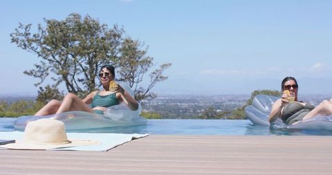 Two women relaxing on clear floats in infinity pool sipping citrus cocktails, city view