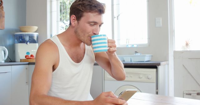 Young man enjoying coffee in a bright, cozy kitchen while browsing his smartphone. Ideal for concepts related to morning routine, lifestyle, technology habits, and casual home activities.