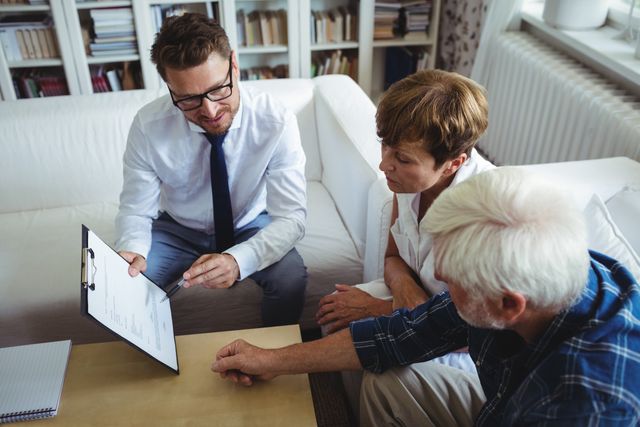 Senior couple consulting with a financial advisor in their living room. The advisor is showing documents on a clipboard, and the couple is attentively listening and discussing their investment plans. Ideal for use in articles or advertisements related to financial planning, retirement advice, senior services, and home consultations.