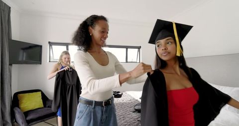 Diverse friends helping graduate into cap and gown in bedroom celebrating milestone