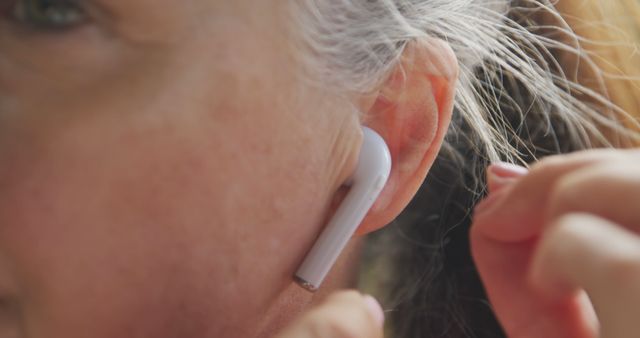 Close-up of a senior woman adjusting white wireless earbuds in ear. Ideal for use in healthcare, technology, or lifestyle contexts to highlight adoption of modern audio devices by elderly people. Suitable for illustrating usage of smart technology, promoting wireless products, or depicting senior citizens embracing tech gadgets.