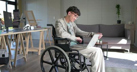 Young man in wheelchair working on laptop in sunlit modern office promoting accessibility