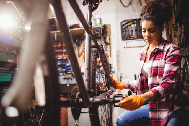 Woman focused on repairing a bicycle in a workshop. Wearing a plaid shirt and orange gloves, illustrating mechanical skills and hands-on work. Ideal for articles about female mechanics, DIY bike maintenance, workspaces, or promoting repair services.
