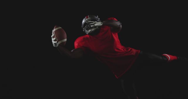 Image shows an American football player in mid-air catching the ball while wearing a helmet and red jersey, isolated on a black background. Can be used for sports event promotions, football training materials, athletic performance illustrations, or advertisements for sports equipment.