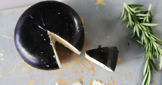 A wheel of black cheese is cut into pieces, with a sprig of rosemary on the side, on a light surface with copy space. Black cheese is a unique culinary item, often infused with edible charcoal for its distinctive color and potential health benefits.