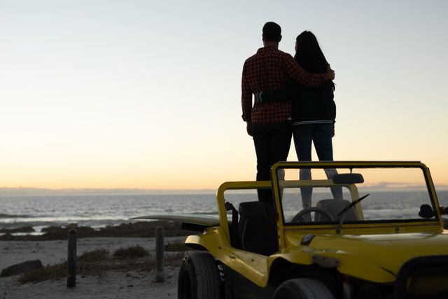 Couple standing by a yellow beach buggy, embracing and admiring the sunset over the sea. Perfect for themes of romance, travel, adventure, and summer holidays. Ideal for use in travel brochures, romantic getaway promotions, and lifestyle blogs.