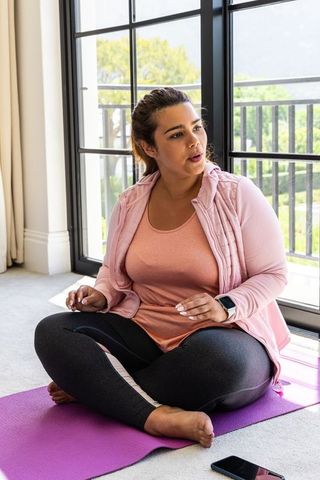 African american woman sitting on yoga mat by balcony glass doors in pink athleisure
