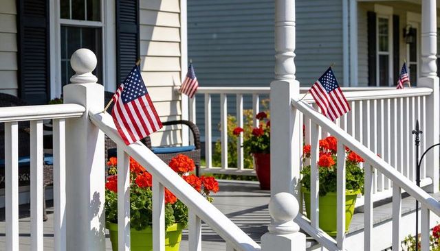 Sunlit american flags decorating white porch railings with red geraniums in lime pots