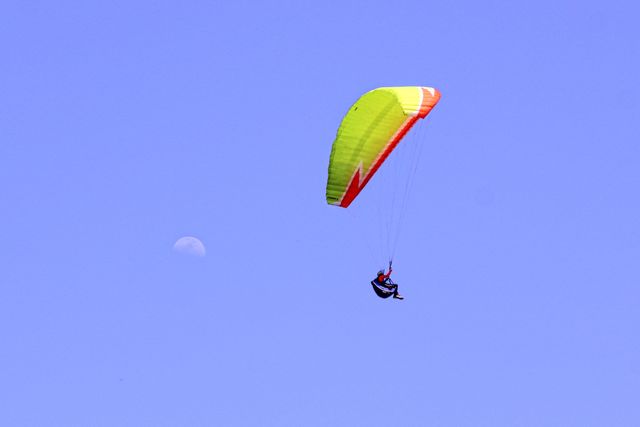 Paraglider soaring gracefully in clear blue sky with half moon visible. Ideal for portraying outdoor adventures, extreme sports, leisure activities, and thrill-seeking experiences. Use in travel promotions, adventure sport articles, or inspirational content.