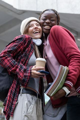 College friends laughing and hugging on campus holding coffee, headphones and textbooks