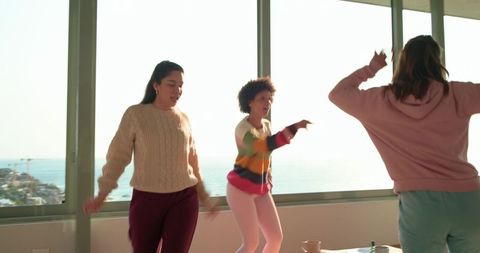 Sunlit multiracial friends dancing in coastal apartment with ocean view and glass walls