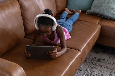Young african american girl listening on headphones using tablet on brown leather sofa