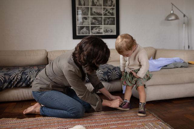 Side view of a young Caucasian mother kneeling on the floor in a sitting room, tying her babys shoe, baby watching her. 