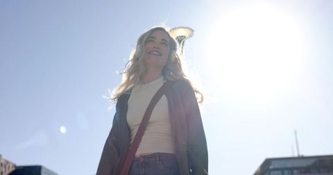 Young woman standing in sunlit urban street wearing headphones, denim, and crossbody bag