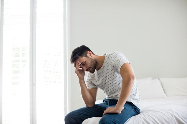 Young man sitting on bed in bright bedroom, looking worried and stressed. Ideal for use in articles or advertisements related to mental health, stress, anxiety, personal reflection, or home life. Can also be used in lifestyle blogs or health and wellness websites.