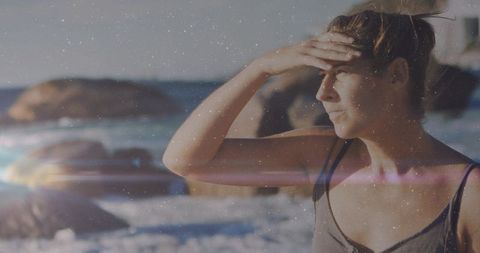 Woman shielding eyes and gazing toward ocean at sunlit rocky beach with lens flare and copy space