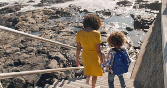 Happy biracial mother and son holding hands walking down steps to beach in the sun, copy space. Motherhood, childhood, togetherness, summer, vacations and free time, unaltered.