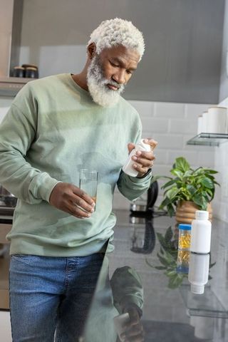 Mature african american man reading pill label holding glass of water in bright kitchen