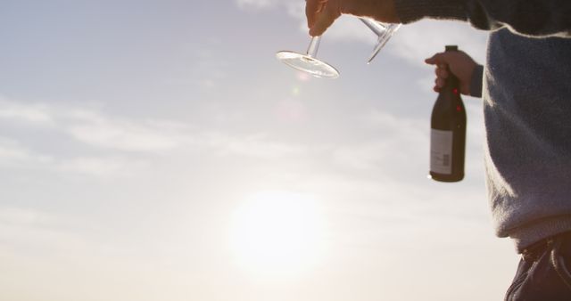 The image captures an individual’s hands holding a wine glass and bottle against a serene sunset sky, creating a relaxed atmosphere. This can be used for content related to leisure activities, celebrations, outdoor events, and lifestyle marketing. The warm lighting enhances a sense of tranquility and enjoyment, making it suitable for advertisements and articles about relaxation, outdoor gatherings, and wine tasting events.