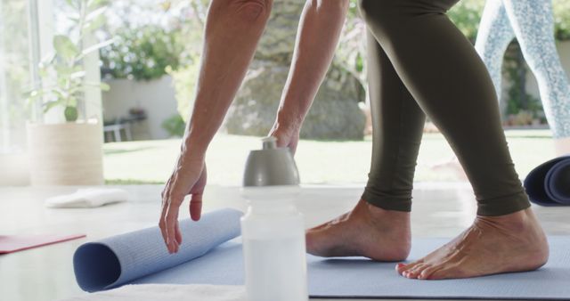 Person rolling up yoga mat after an outdoor workout session with water bottle nearby. Great for illustrating healthy lifestyle, fitness routines, mindfulness practices, yoga activities, and home exercise environments.
