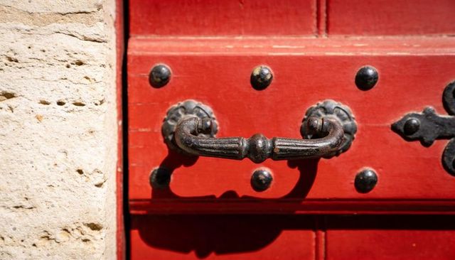 Ornate cast-iron door pull casting shadow on weathered red wooden door with black studs
