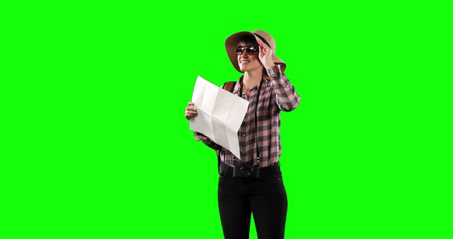 Woman wearing hat and sunglasses is holding map, positioned against green chroma key background. Excellent for travel-related themes, video editing, digital content, and adventure promotion.