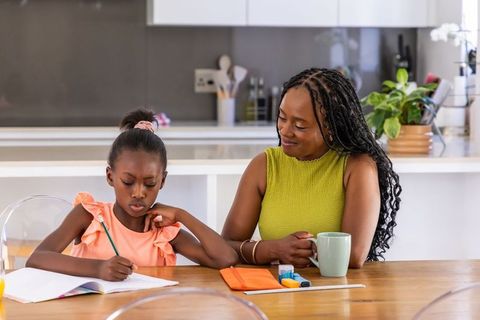 African american mother guiding daughter with homework at bright kitchen table with coffee