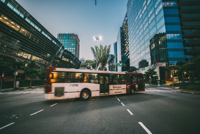 Bus passing through a city intersection at twilight, surrounded by modern buildings. This image can be used for themes related to urban living, public transportation, city infrastructure, and evening commutes.