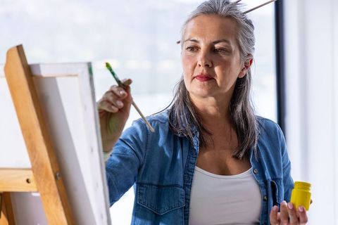 Mature woman painting on canvas in sunlit home studio holding brush and yellow paint jar