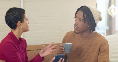 Multicultural friends talking on couch while woman shows phone and man holds mug