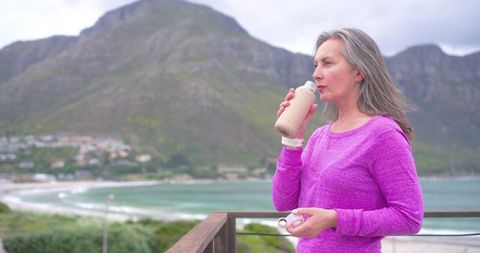 Mature woman drinking from reusable bottle on coastal balcony overlooking bay