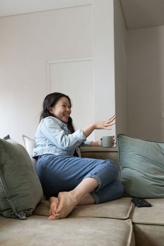 Asian woman relaxing on sofa holding mug and smiling in minimalist neutral living room