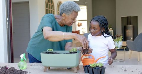 African grandma teaching granddaughter potting seedlings in planter with orange watering can