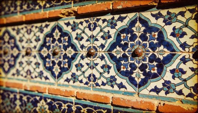 Glazed ceramic tile band with blue arabesque floral medallions and aged metal studs on brick facade