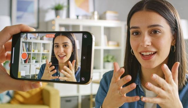 Young woman recording lifestyle vlog on smartphone in cozy home office, gesturing to camera