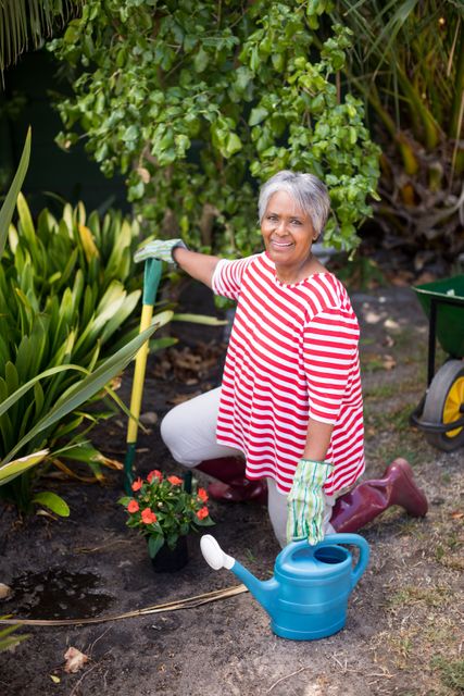 Senior woman kneeling in yard while gardening, holding a shovel and surrounded by plants and flowers. She is wearing a red and white striped shirt, gloves, and red boots. A blue watering can and a potted flower are nearby. Ideal for use in articles about gardening, healthy lifestyles for seniors, outdoor hobbies, and nature activities.