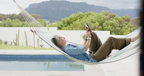 Mature woman relaxing in hammock by modern pool reading e-reader with mountain view