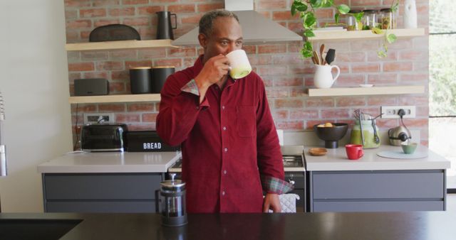 Man stands in modern kitchen with brick wall, wooden shelves, and various kitchen items, drinking coffee. Useful for concepts related to home, lifestyle, morning routines, kitchen design, and casual moments.