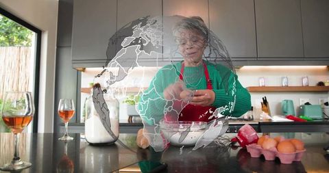 Senior woman cooking in modern kitchen cracking eggs into bowl wearing red apron and green sweater