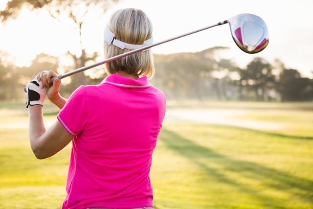 This image shows a female golfer in a pink shirt swinging a golf club on a sunny day. Ideal for use in sports and fitness articles, golfing advertisements, outdoor activity promotions, and healthy lifestyle campaigns.